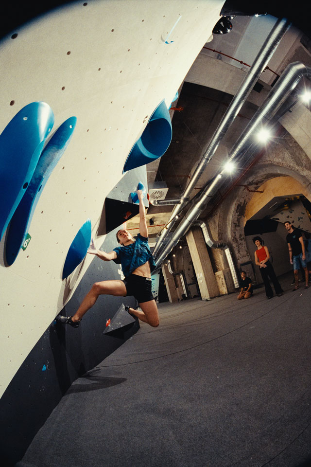 Boulderin hält sich an blauen Griffen fest an einem Überhang im urban apes Fhain Berlin. Drei Freunde schauen zu.