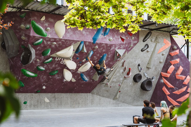 Person klettert an Outdoor-Boulderwand von urban apes bright site Berlin, umgeben von Zuschauern und grünen Blättern.