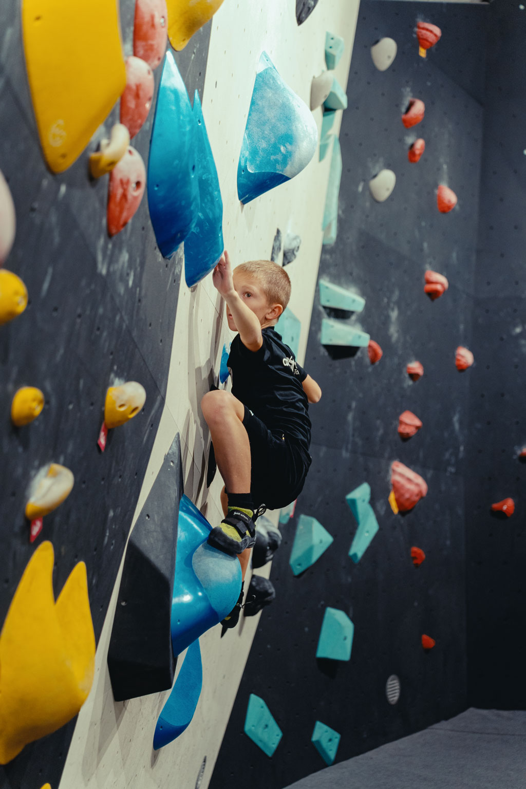 Junge greift nach Griff an Boulderwand, voller Konzentration beim Klettern.